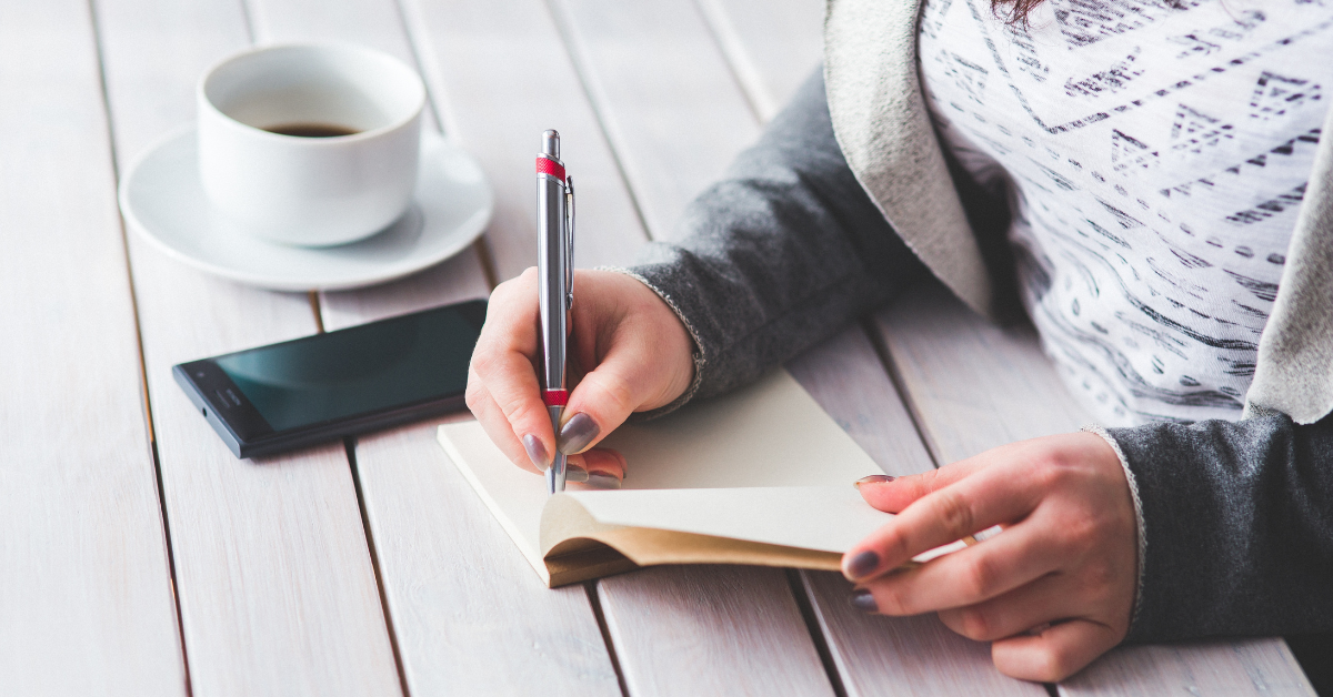 A woman's hands journaling in a notebook with a cup of coffee beside her, symbolizing self-care during perimenopause and menopause.