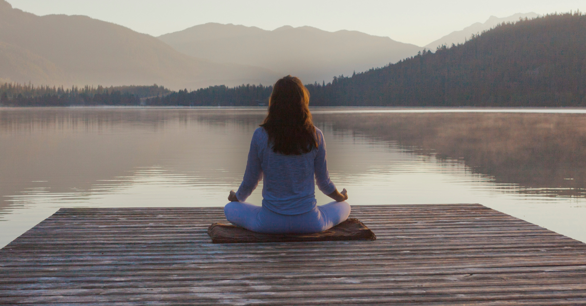 Midlife woman sitting cross-legged and meditating by a calm lake, reflecting a peaceful and supportive approach to hormone health and healing